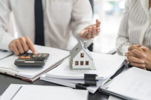 A property manager talking to a property owner working out finances on a calculator with a wooden house figure on top of the documents.